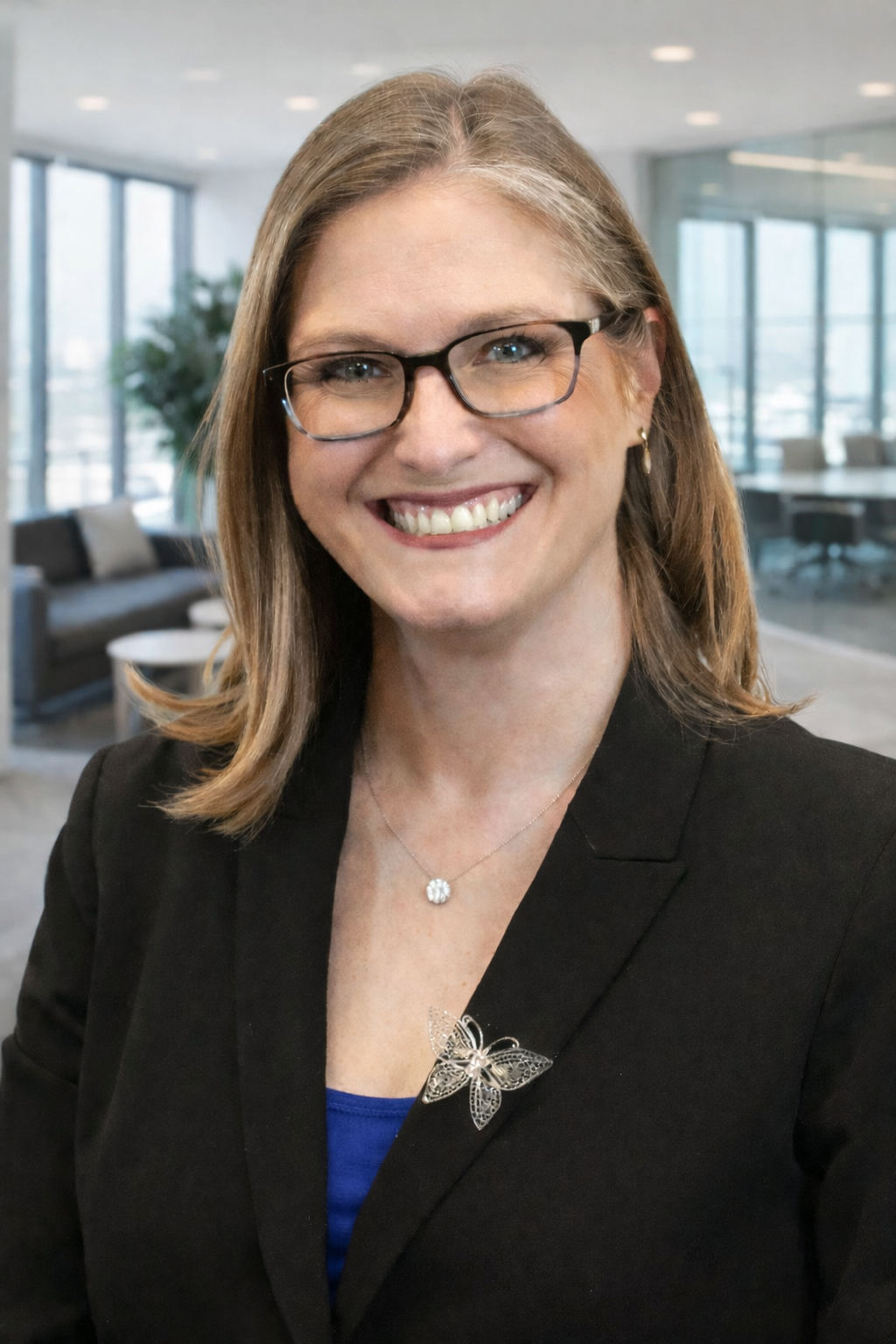 Smiling woman with glasses in a black blazer and blue shirt, butterfly brooch, in a modern office setting.