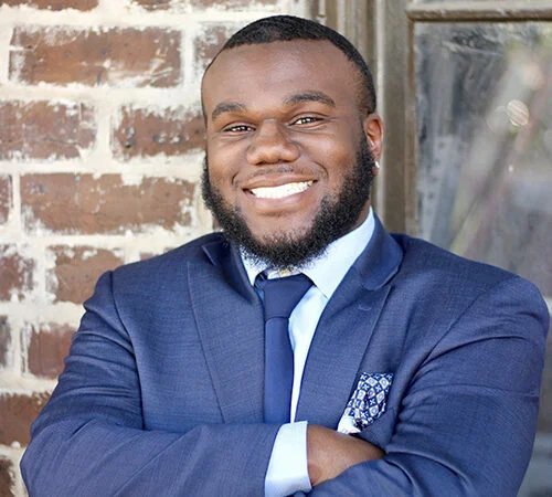 Smiling man in a navy suit with a light blue shirt and patterned pocket square, standing against a brick wall.