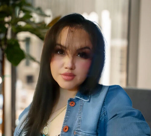 Young woman with long dark hair wearing a denim jacket, sitting indoors with a plant in the background.