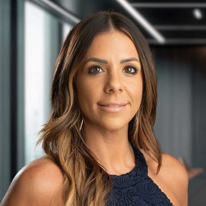 Woman with long brown hair wearing a navy blue top, smiling in a modern office setting with large windows.