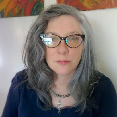 Woman with long gray hair and cat-eye glasses, wearing a dark top, against a light background with colorful artwork.