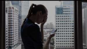 Woman in a dark shirt stands by a window, holding a phone and covering her face, with city buildings in the background.