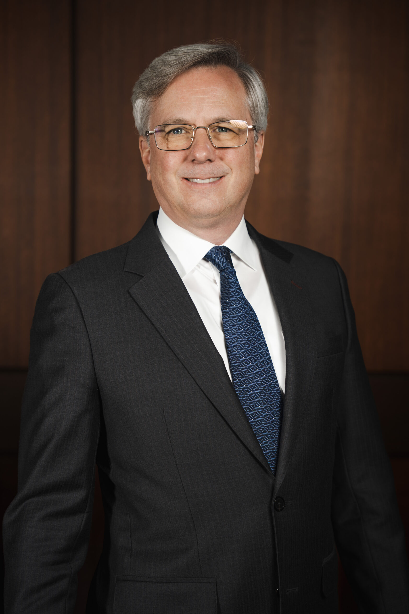 Man in a dark suit and blue patterned tie smiles against a wooden background.