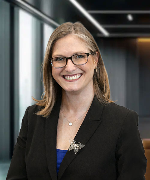 Smiling woman with glasses, wearing a black blazer and blue top, stands in a modern office setting.
