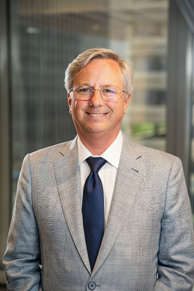Smiling man in a light gray suit and navy tie, standing in an office with large windows in the background.
