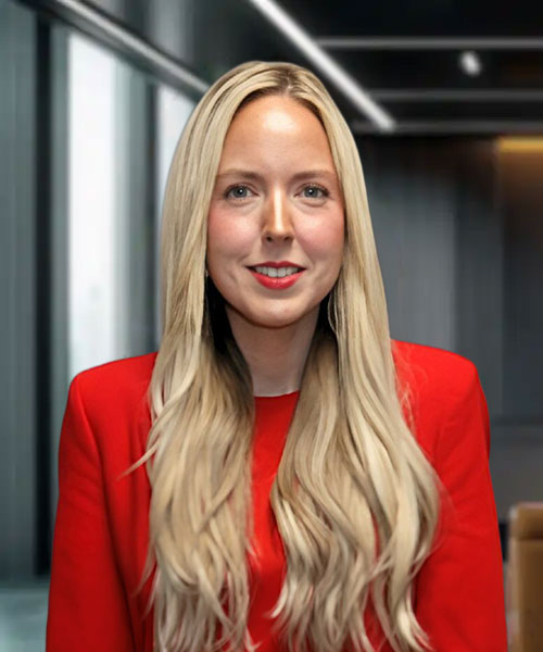 Woman with long blonde hair wearing a red blazer, smiling in a modern office setting with soft lighting.