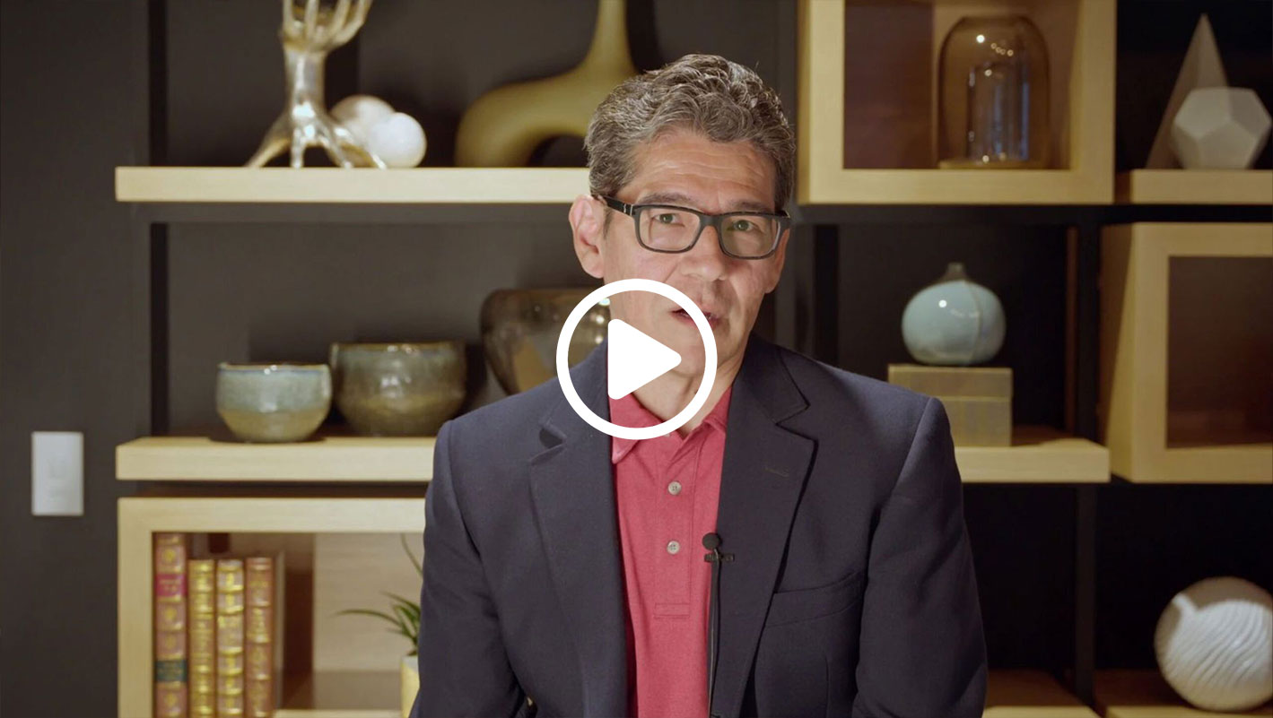 Man in a blazer and red shirt speaks in front of a shelf with decorative items and books. Play button overlay.