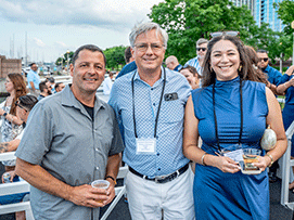 Three people pose together outdoors at a social event, smiling and holding drinks, with a marina in the background.