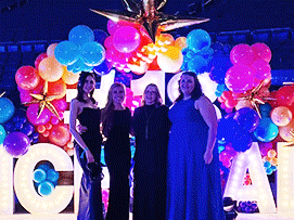 Four women in elegant dresses pose in front of a colorful balloon arch with bright pink, blue, and gold accents.