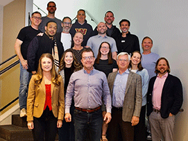 Group photo of 18 people on stairs, smiling, wearing casual and business attire, with a light background.