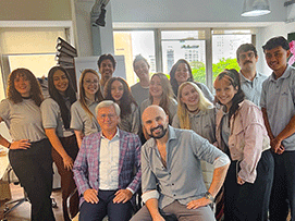 Group photo of a diverse team in a bright office, featuring 13 people in casual attire, with two seated in front.