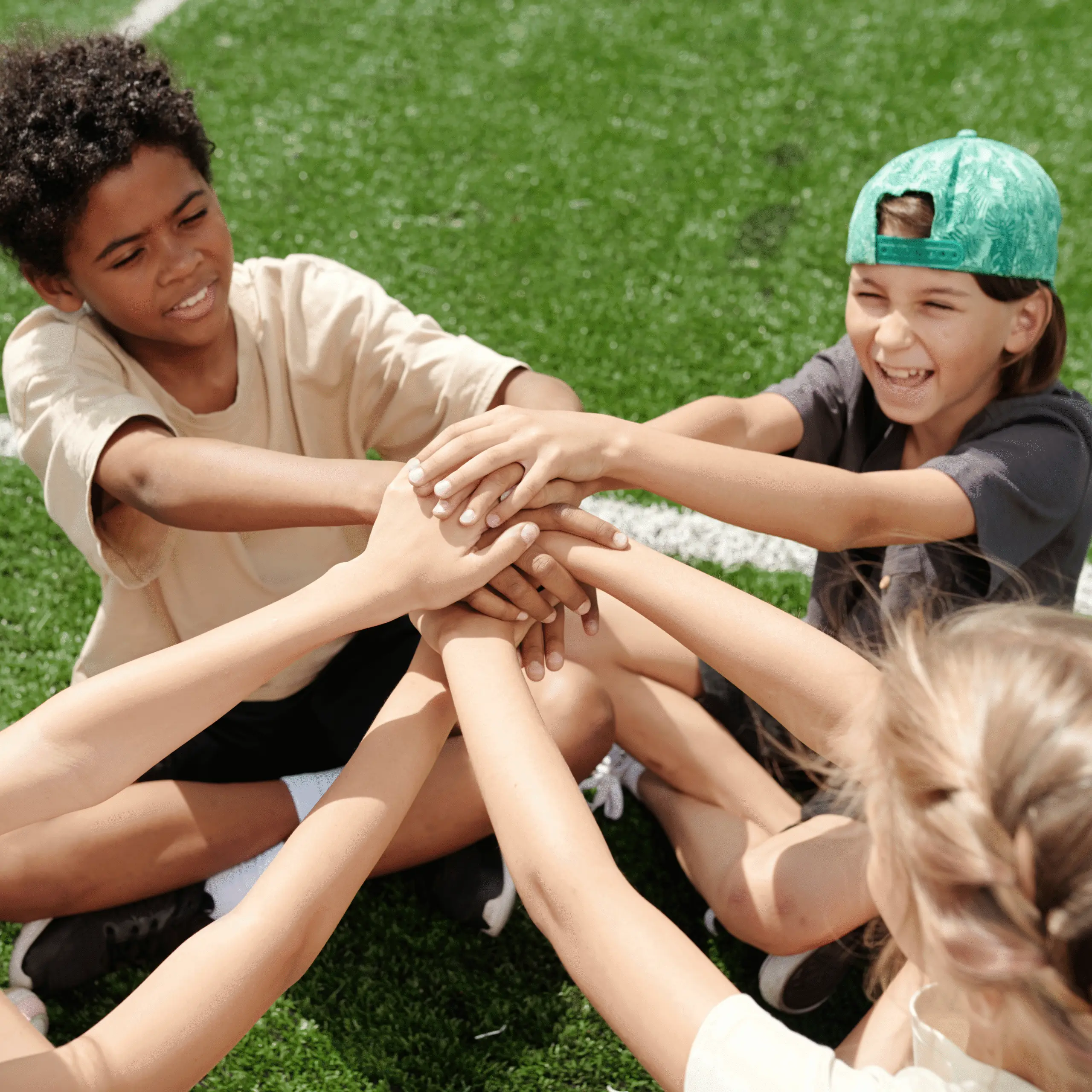Children sitting on green turf, stacking hands together in a circle, smiling and engaged in teamwork.