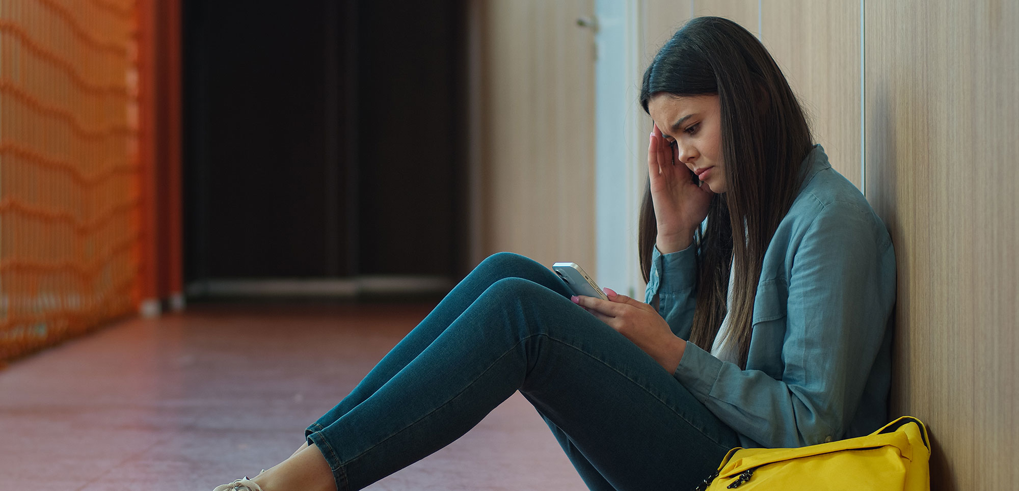 Young woman sitting against a wall, looking stressed while using her phone, with a yellow bag beside her.