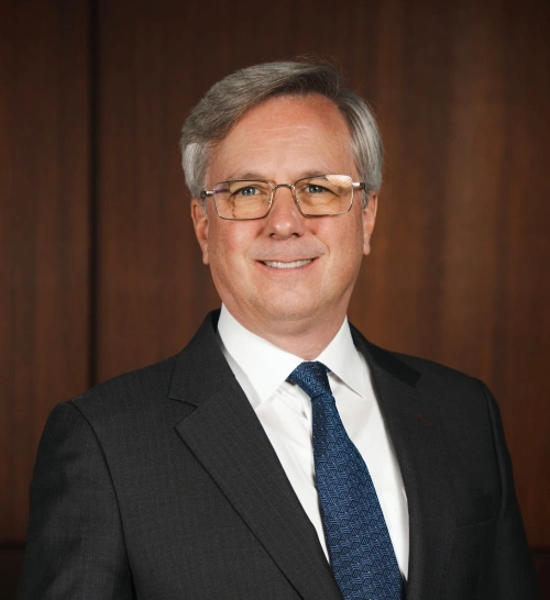 Man in a dark suit and blue tie, smiling against a wooden background. He has gray hair and glasses.