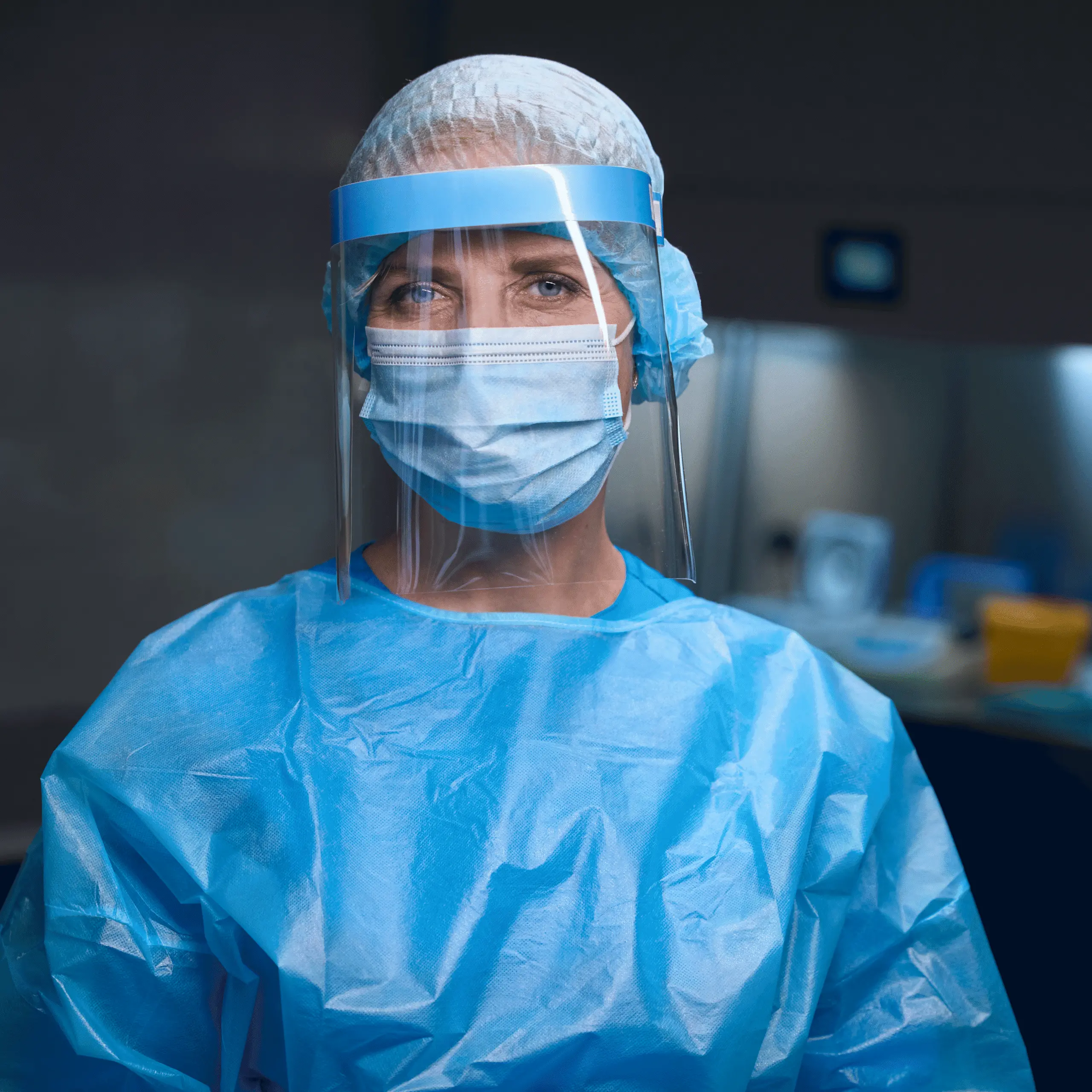 Healthcare worker in blue gown, mask, and face shield, standing in a clinical setting.