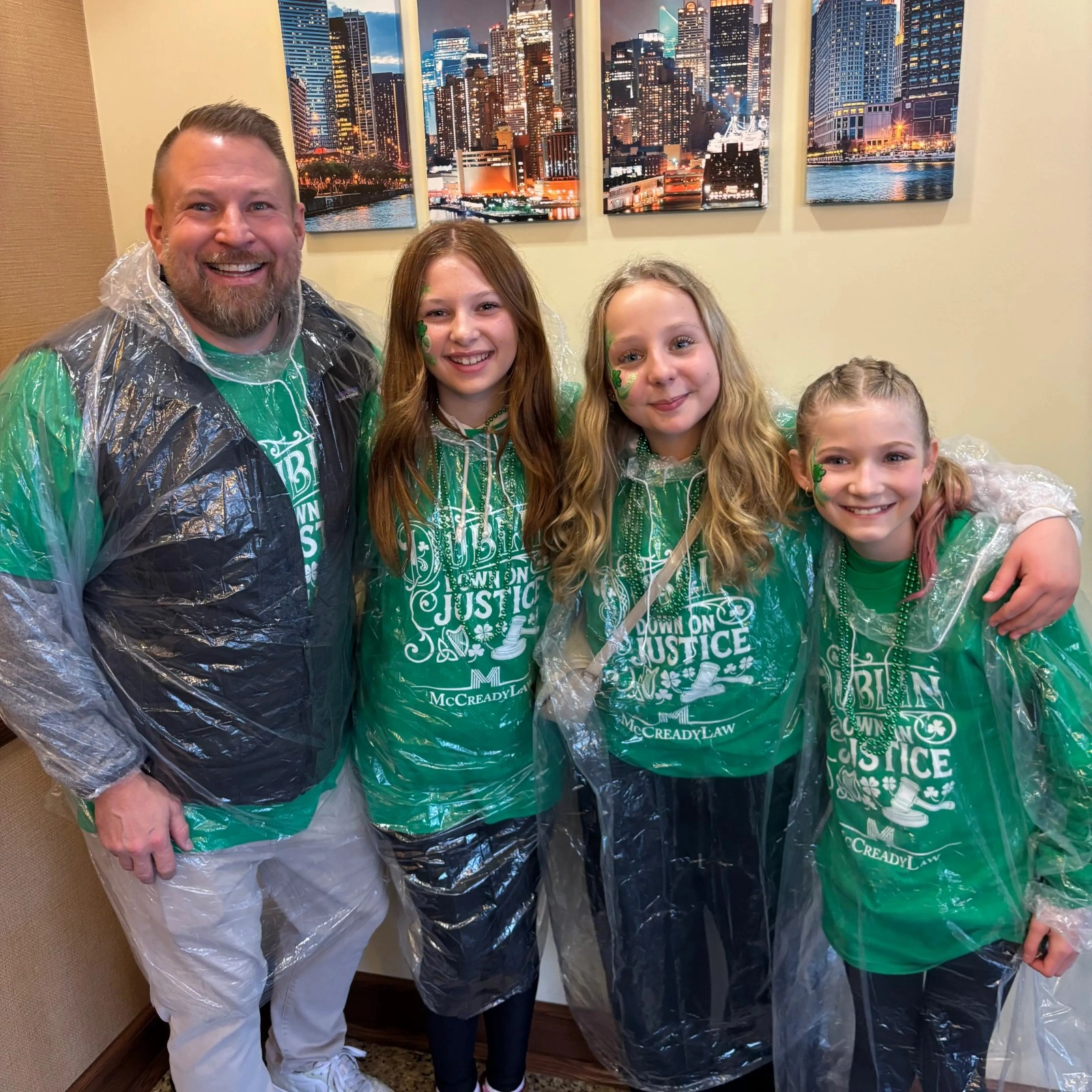 Four people in green shirts with "Countdown on Justice" text, wearing clear ponchos, smiling in front of a city skyline.