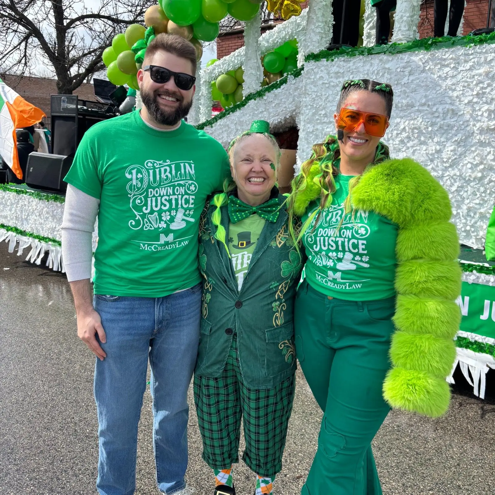 Three people in green outfits celebrate St. Patrick's Day, smiling in front of a decorated float with balloons.