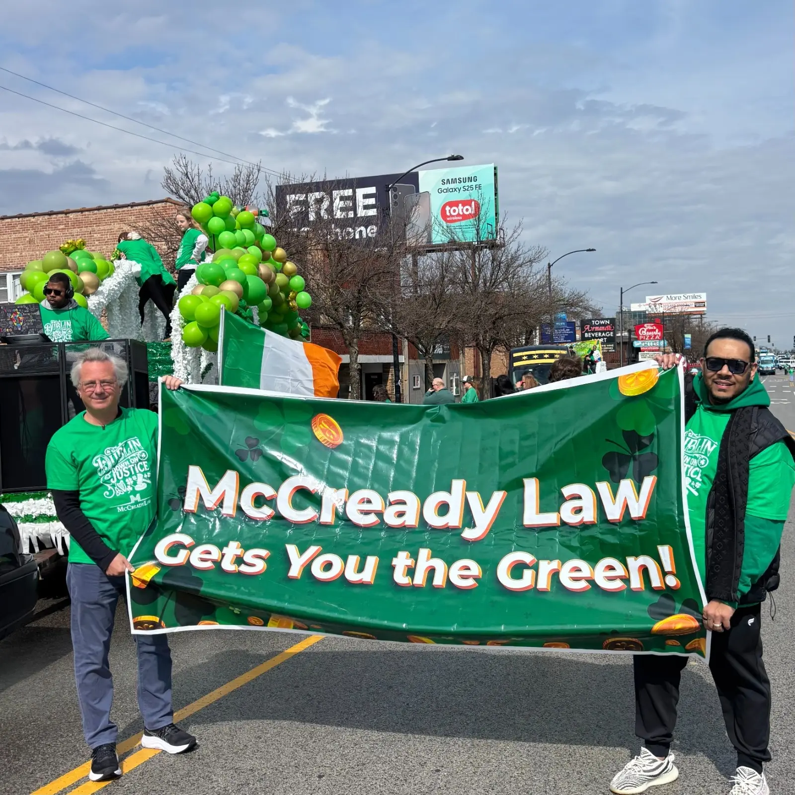 Two men hold a green banner for McCready Law at a parade, with an Irish flag and festive balloons in the background.