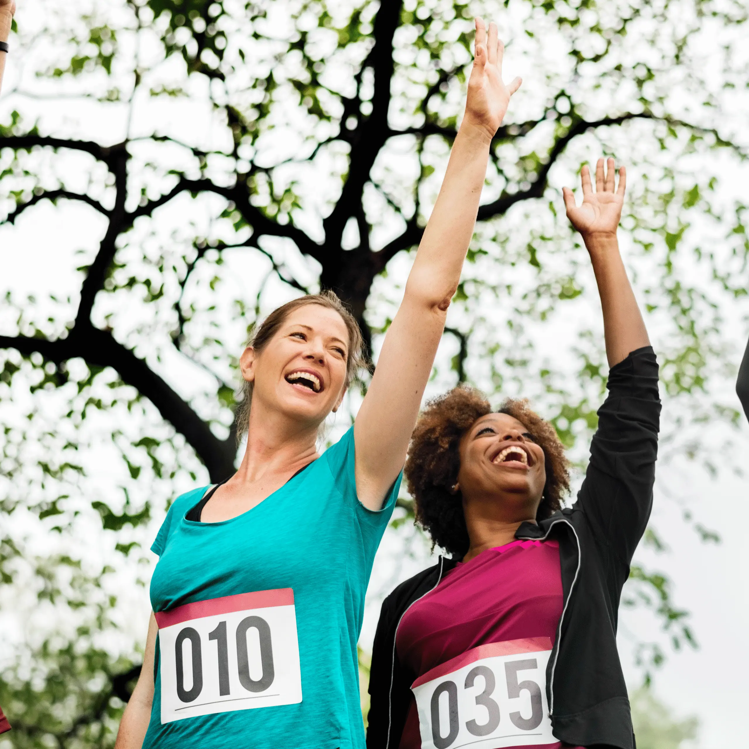 Two women celebrate with raised arms, wearing race bibs 010 and 035, under a leafy tree in a park.
