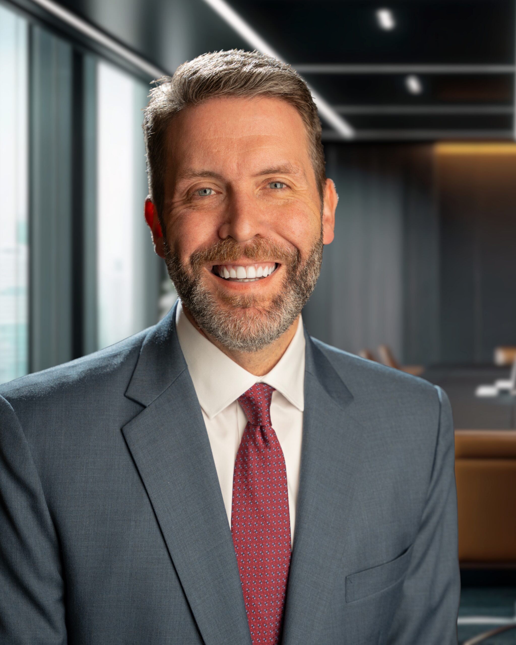 Smiling man in a blue suit and red tie, standing in a modern office with large windows and a conference table.