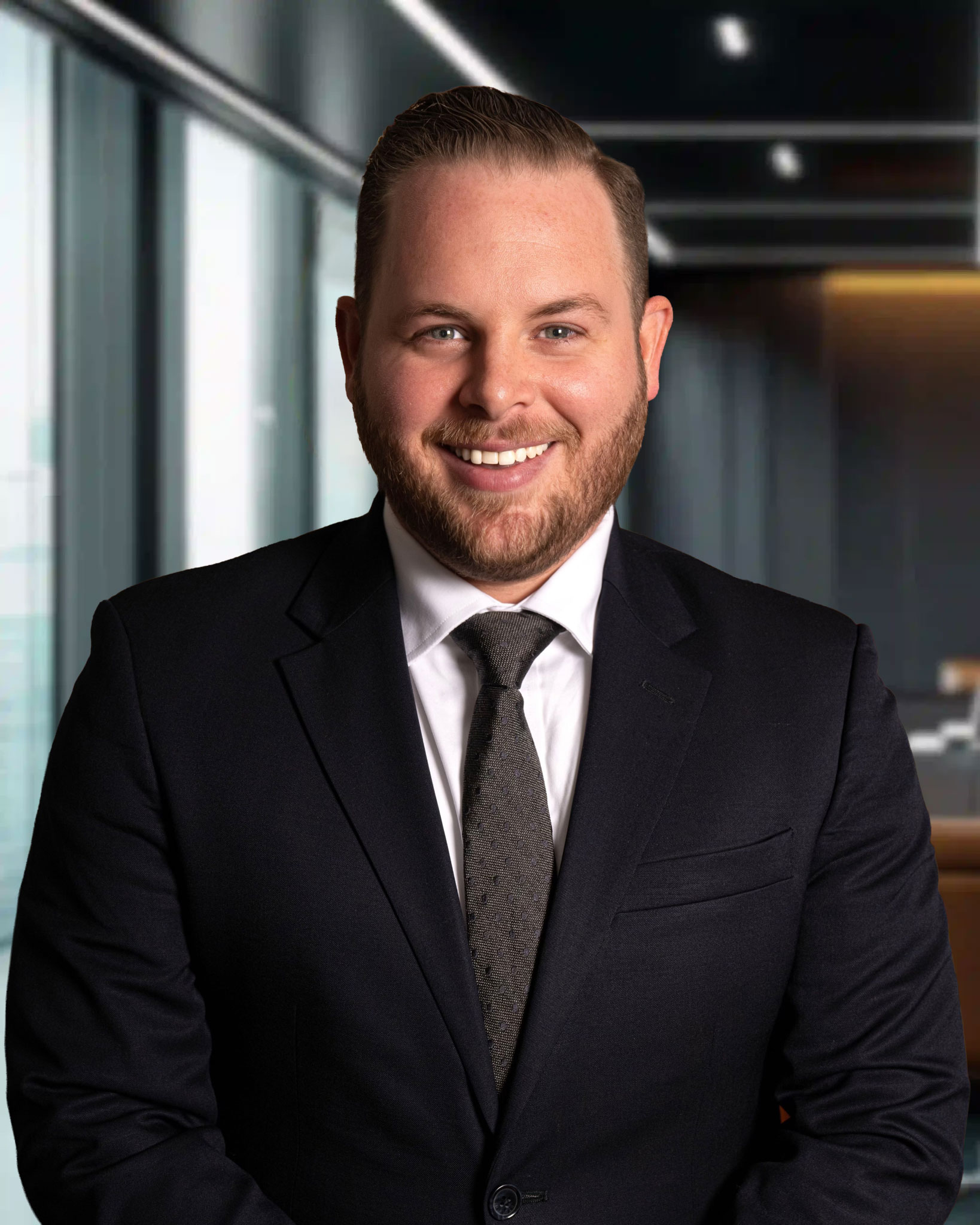Smiling man in a black suit and tie, standing in a modern office with glass windows and soft lighting.