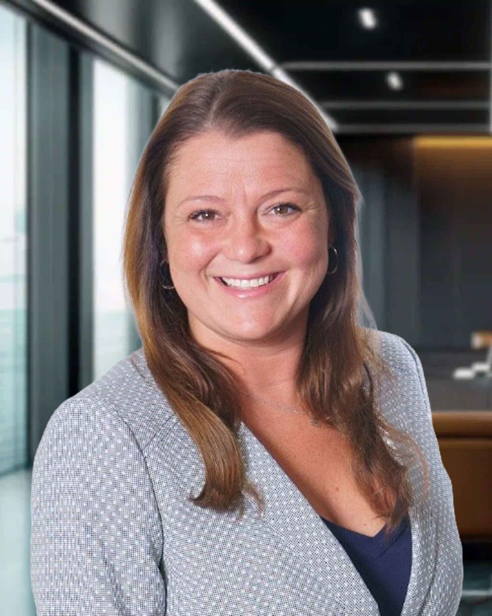 Smiling woman with long brown hair in a gray blazer, standing in a modern office with large windows.