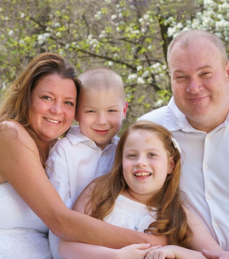 Smiling family of four in white clothing, surrounded by blooming trees in a sunny outdoor setting.