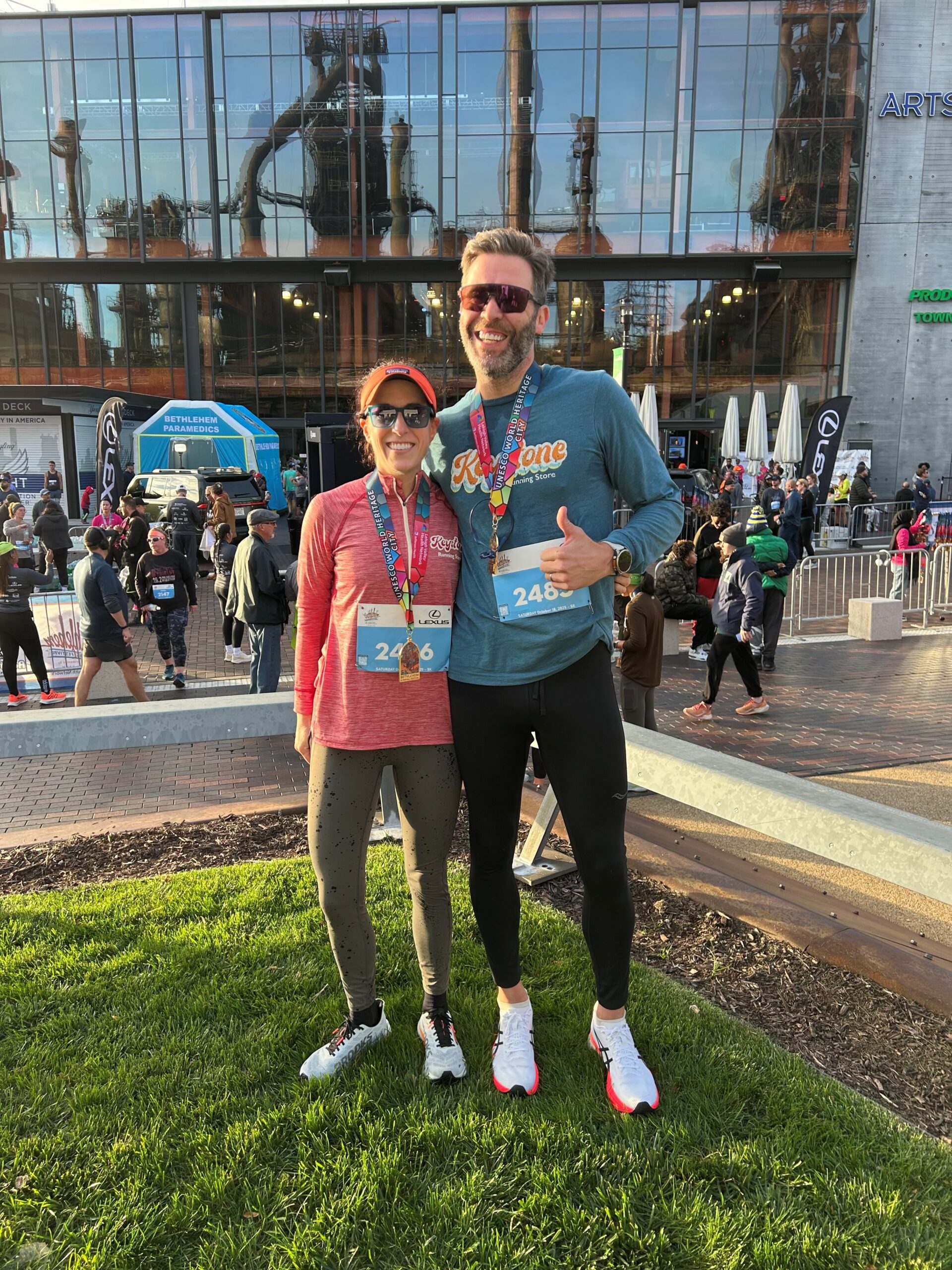 Two runners pose together, smiling, wearing medals and race bibs, with an industrial building in the background.