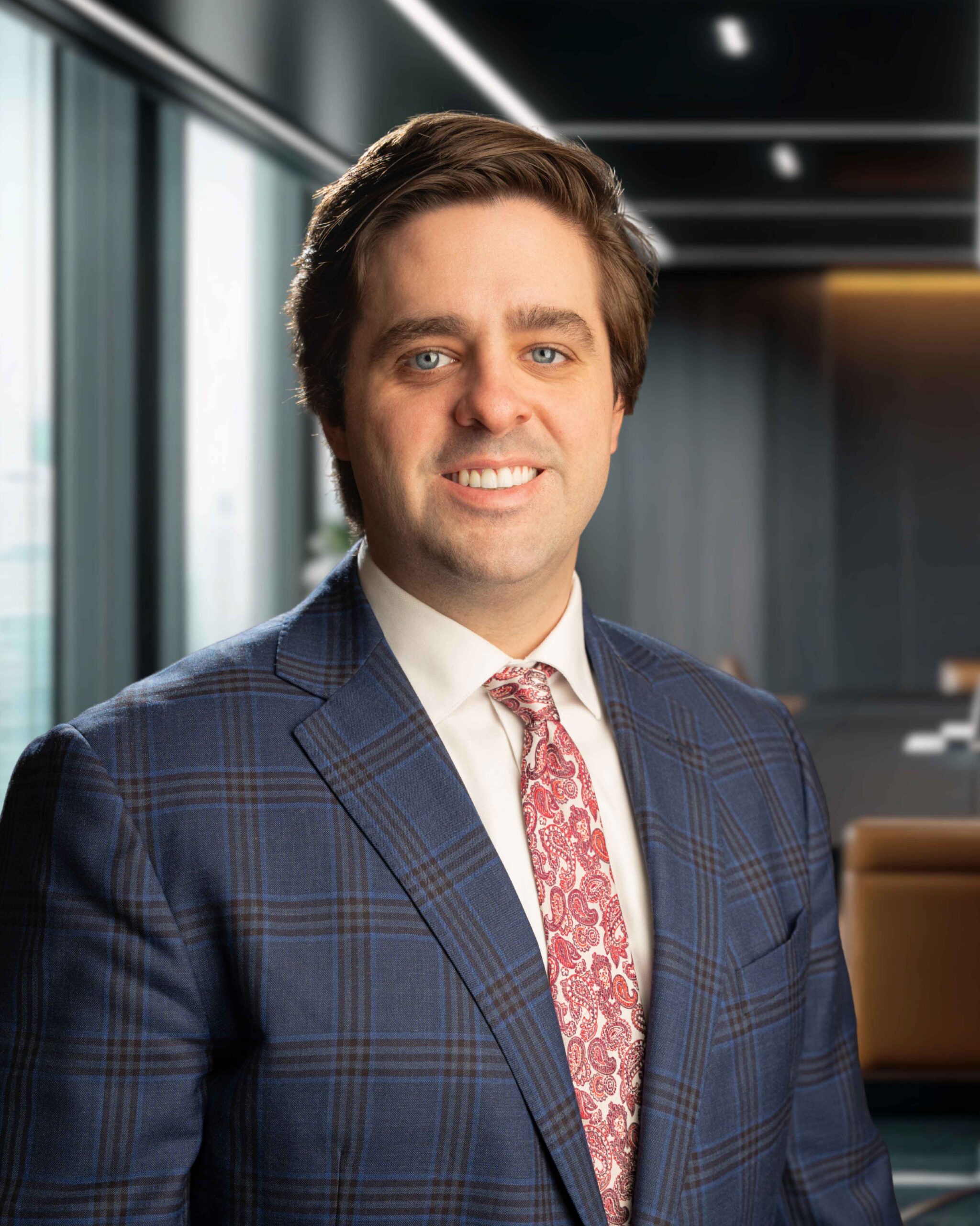 Man in a blue plaid suit and red paisley tie smiles in a modern office setting with large windows.