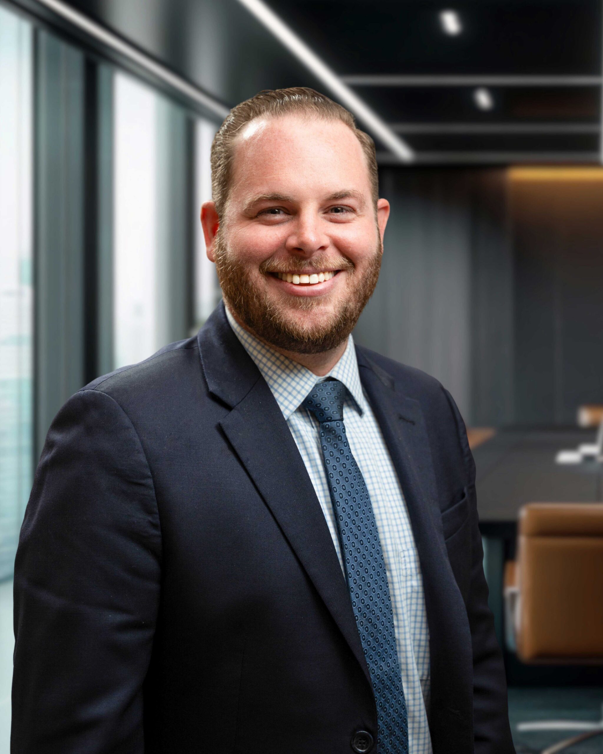 Smiling man in a suit with a patterned tie, standing in a modern office with large windows and a conference table.