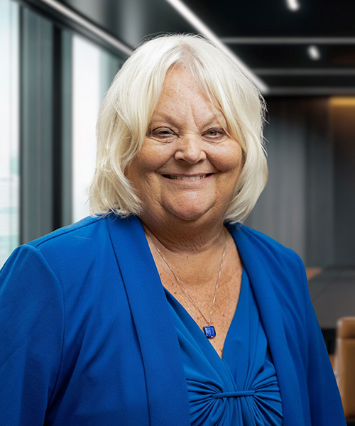 Smiling woman with shoulder-length blonde hair, wearing a blue blazer, in a modern office setting.