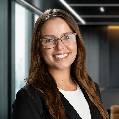 Smiling woman with long brown hair and glasses, wearing a black blazer, in a modern office setting.